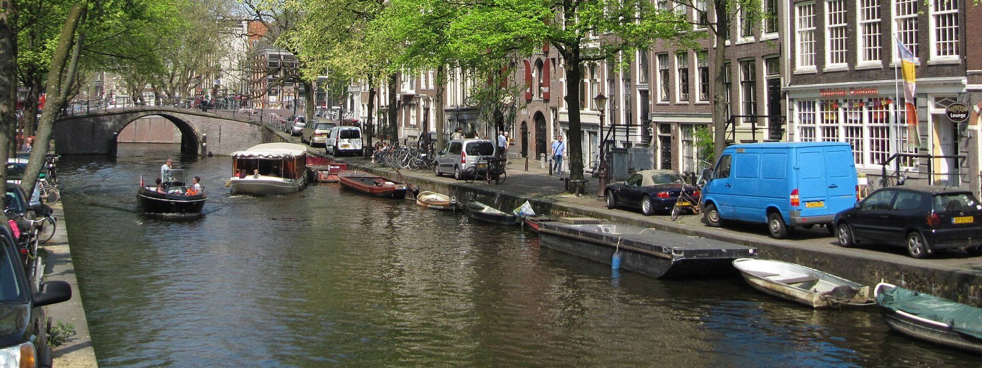 Boats and houseboats moored along an Amsterdam canal, with a stone arched bridge and gabled canal houses behind