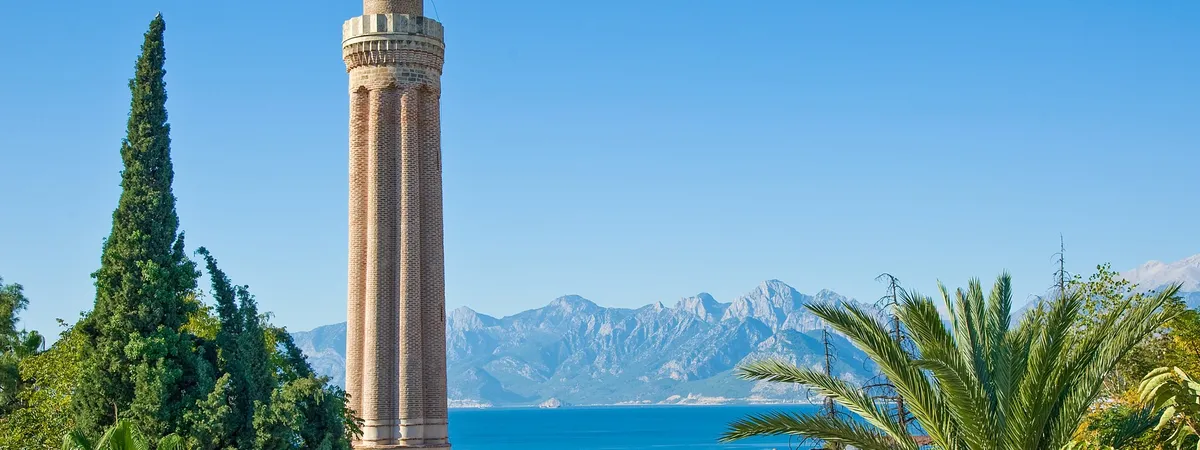 The fluted brick minaret of the Yivli Minare mosque rising above palm trees, with the Mediterranean Sea and the Taurus Mountains behind it