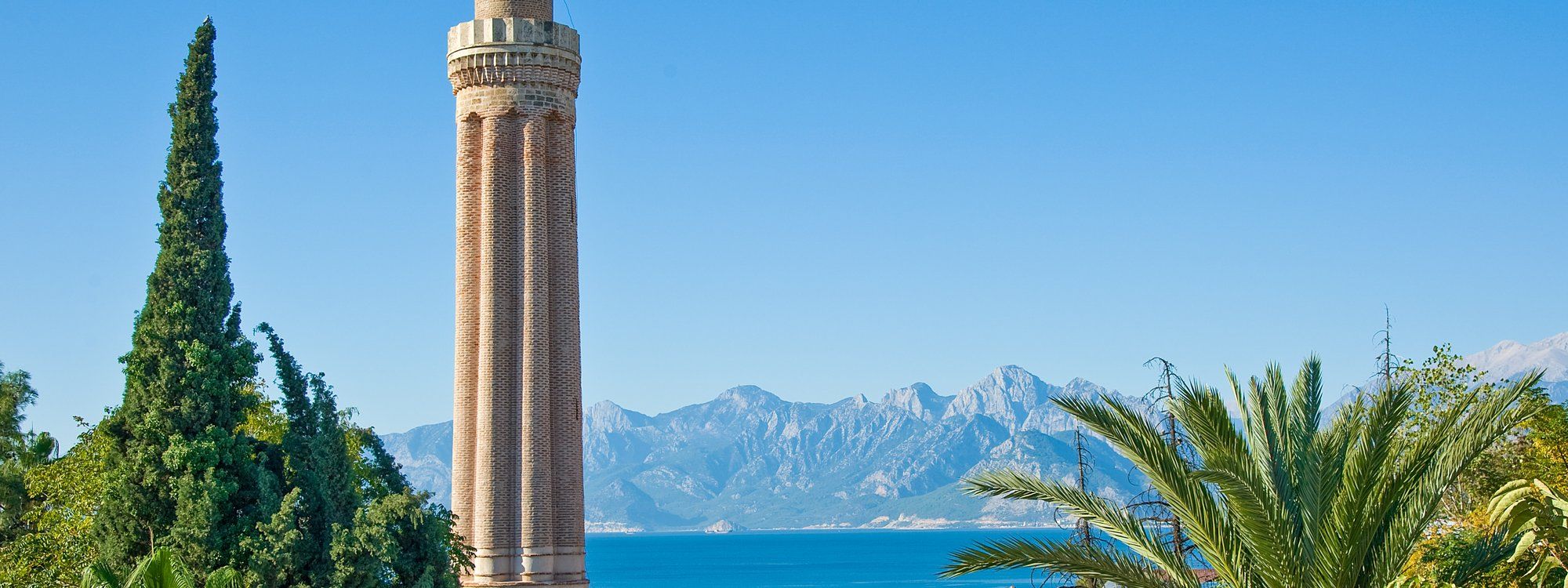 The fluted brick minaret of the Yivli Minare mosque rising above palm trees, with the Mediterranean Sea and the Taurus Mountains behind it
