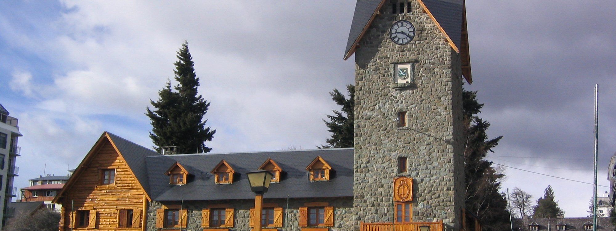 The stone clock tower and Alpine-style municipal building of Bariloche's Centro Cívico, seen across the plaza with pine trees around it