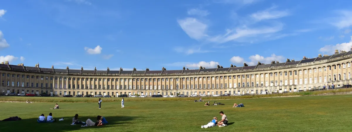 The full sweep of the Royal Crescent in Bath, its curved Georgian terrace facing a wide lawn under a blue sky