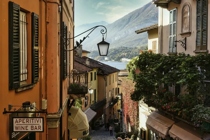 A narrow stepped lane in Bellagio between shuttered houses, opening onto Lake Como and the mountains behind