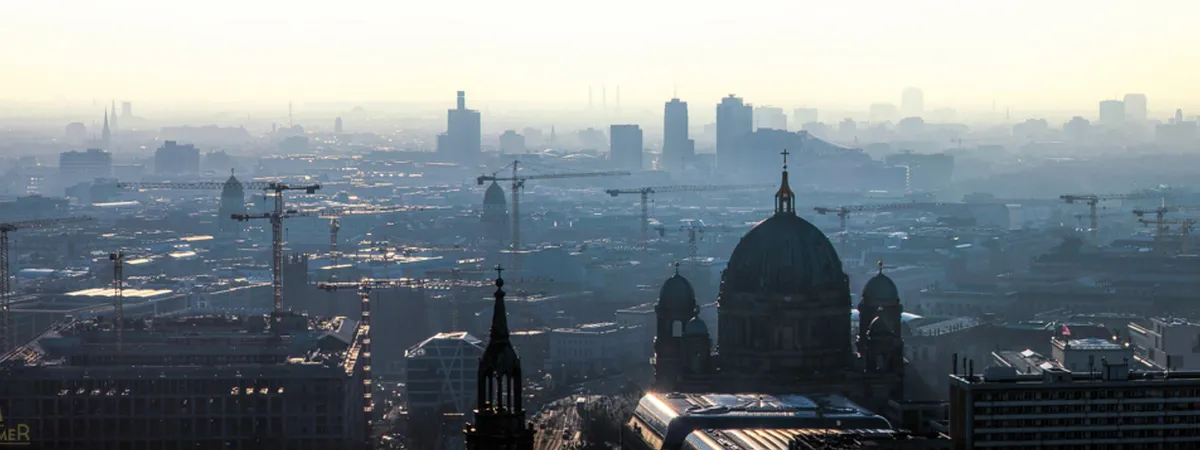 Hazy dawn view over the Berlin skyline from a rooftop, with the dome of Berlin Cathedral in the foreground and construction cranes across the city