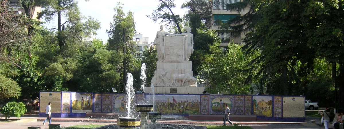 El mural de mosaico de azulejos españoles y la fuente de la Plaza España, con un monumento de piedra y árboles detrás, en el centro de Mendoza