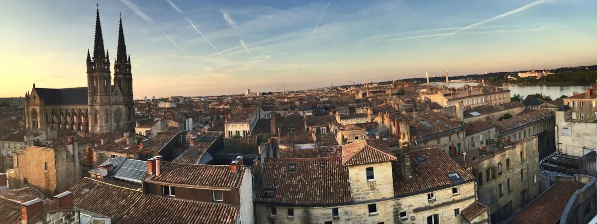 Rooftop view over the Chartrons district of Bordeaux at sunset, with the twin Gothic spires of the Église Saint-Louis-des-Chartrons rising above red-tiled roofs toward the Garonne river