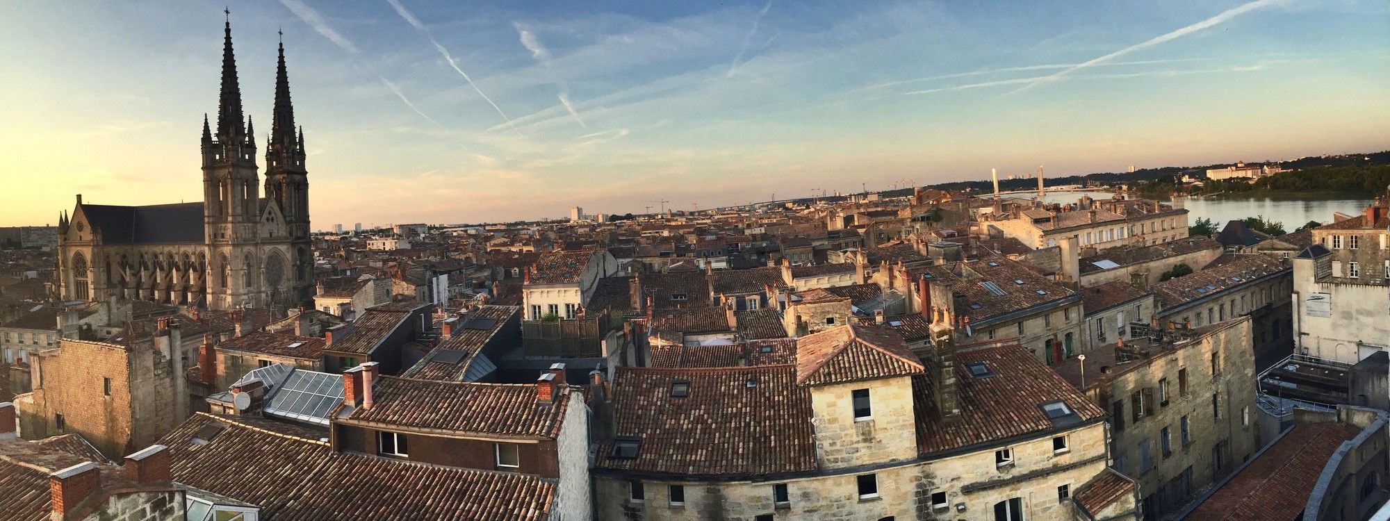 Rooftop view over the Chartrons district of Bordeaux at sunset, with the twin Gothic spires of the Église Saint-Louis-des-Chartrons rising above red-tiled roofs toward the Garonne river