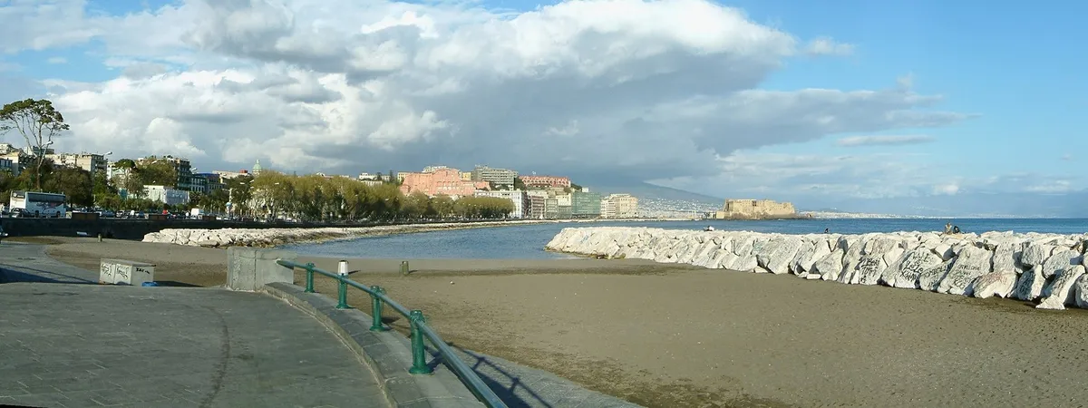 The Naples waterfront seen from Via Caracciolo, with the city skyline and Castel dell'Ovo along the bay and the silhouette of Mount Vesuvius in the distance