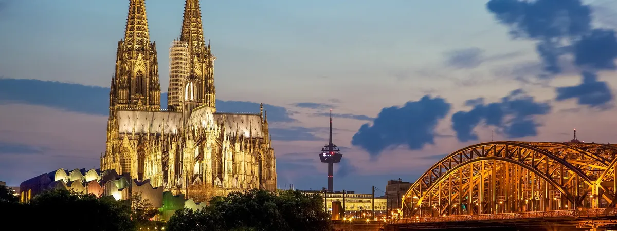 Cologne Cathedral's twin spires lit up at dusk, with the golden trusses of the Hohenzollern Bridge crossing the Rhine in the foreground