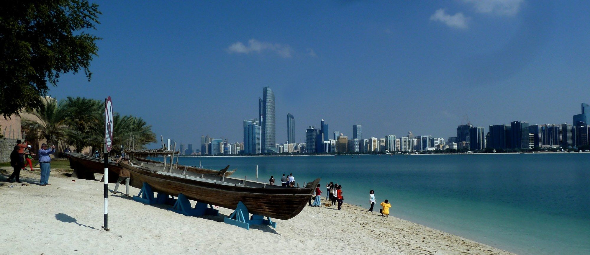Traditional wooden dhow boats on the sandy Corniche beach, with the Abu Dhabi skyline including Etihad Towers across the water