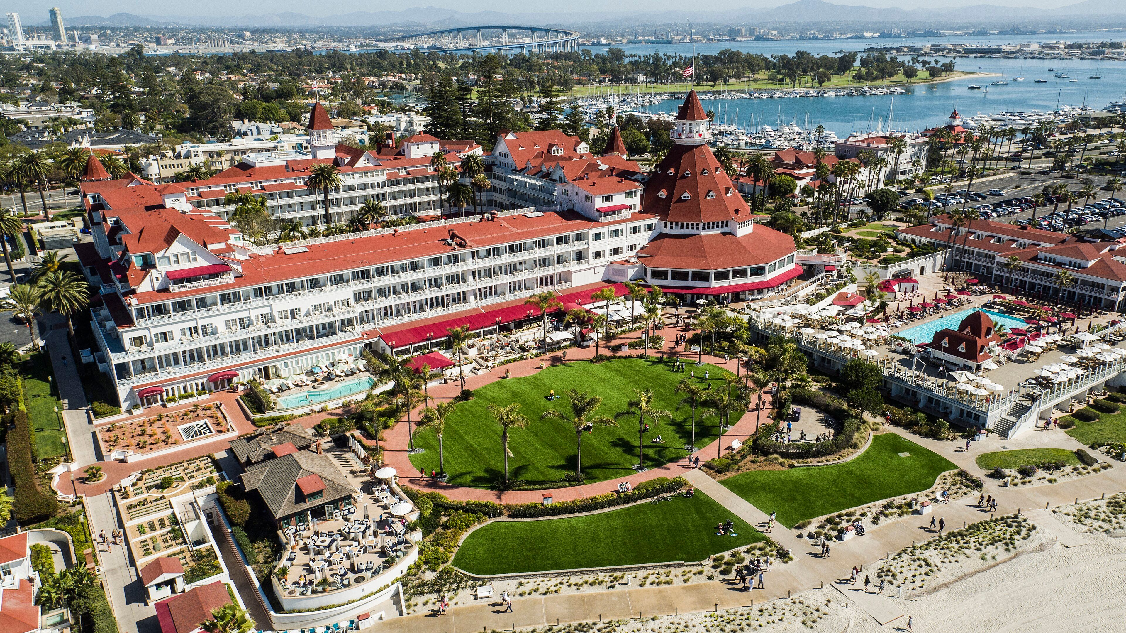 Aerial view of the red-roofed Victorian Hotel del Coronado with San Diego Bay and the Coronado Bridge in the background