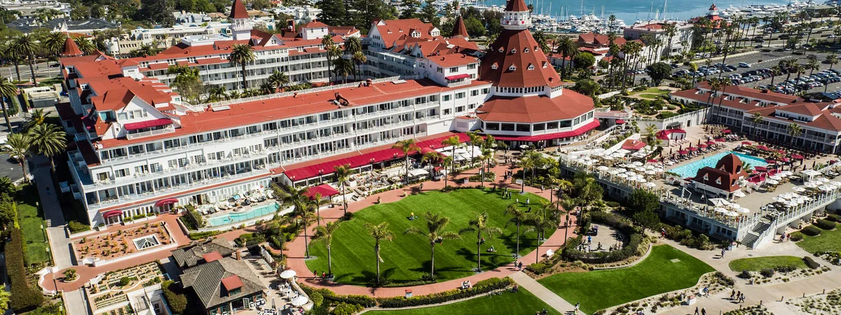 Aerial view of the red-roofed Victorian Hotel del Coronado with San Diego Bay and the Coronado Bridge in the background