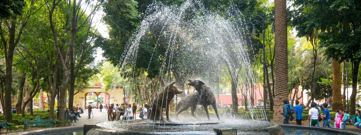 The bronze Fuente de los Coyotes fountain in the Jardín Centenario, Coyoacán, surrounded by trees and visitors
