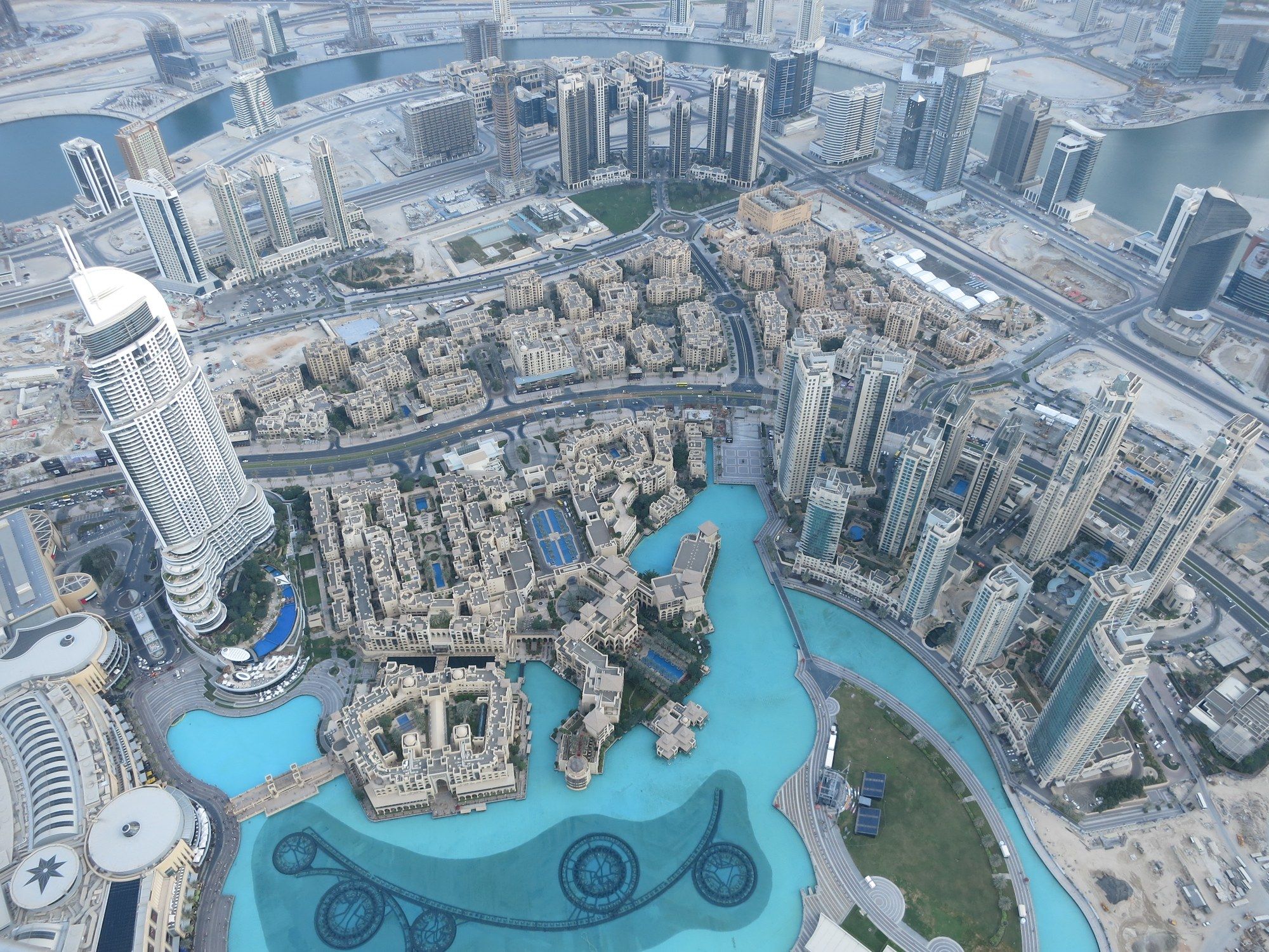 Aerial view down onto the Dubai Fountain lake and the Souk Al Bahar complex, with the Burj Khalifa's shadow stretching across the water