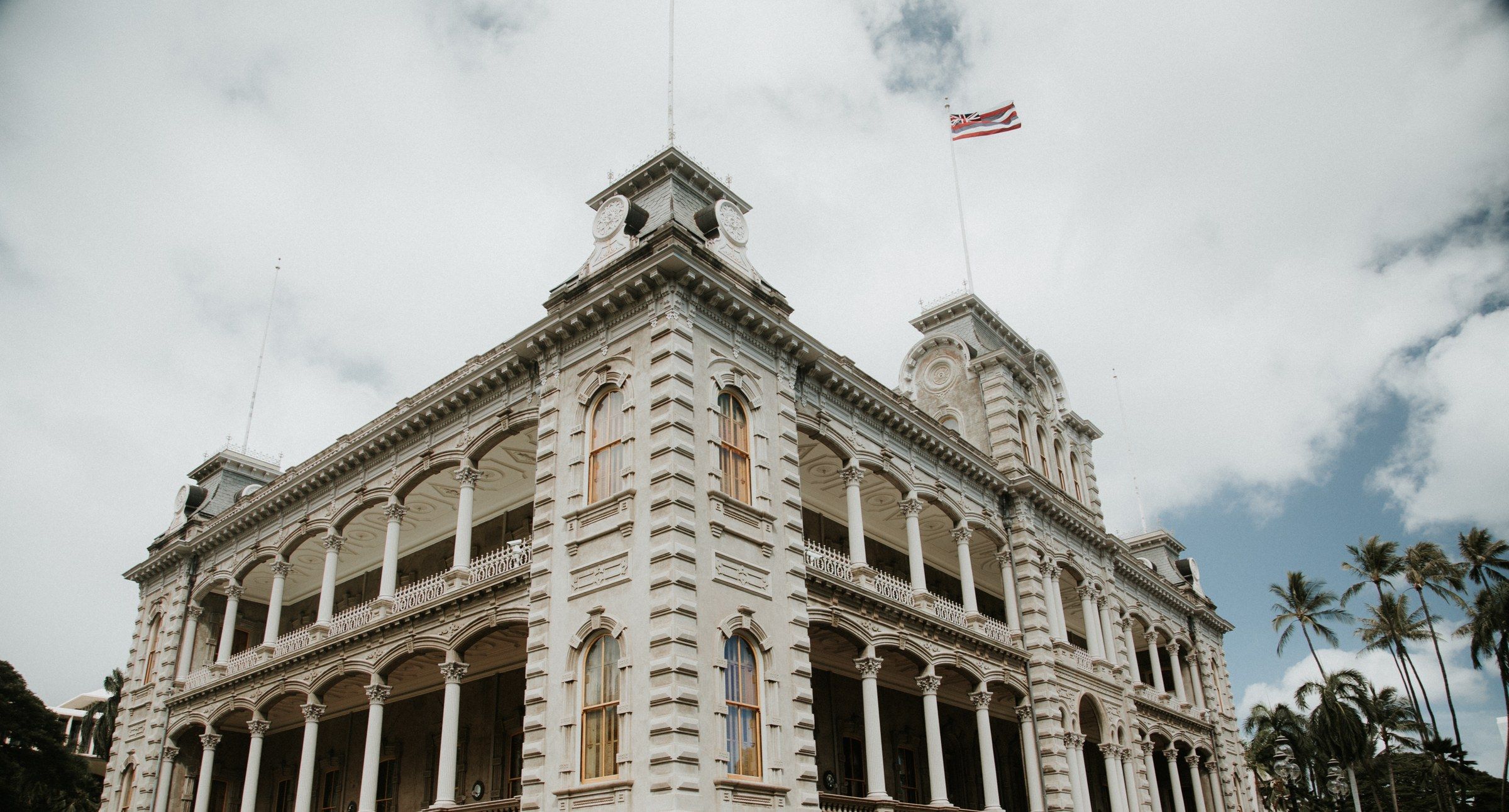 The ornate Renaissance-style facade of ʻIolani Palace, the only royal palace on U.S. soil, with the Hawaiian flag flying above and palm trees beside it