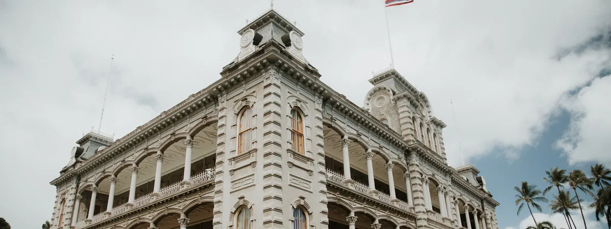 The ornate Renaissance-style facade of ʻIolani Palace, the only royal palace on U.S. soil, with the Hawaiian flag flying above and palm trees beside it