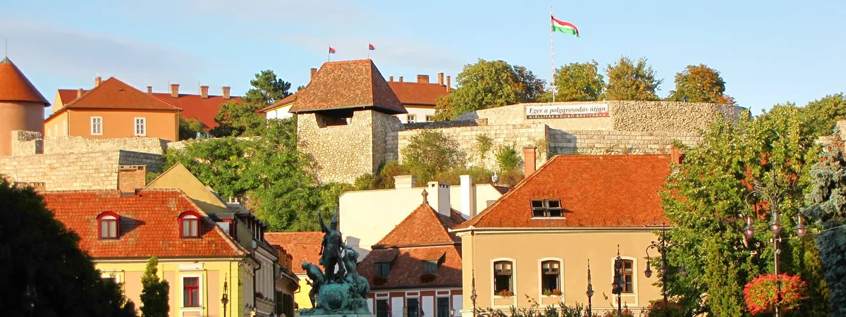 Eger Castle's stone ramparts and a Hungarian flag above the old town's red-tiled rooftops at sunset