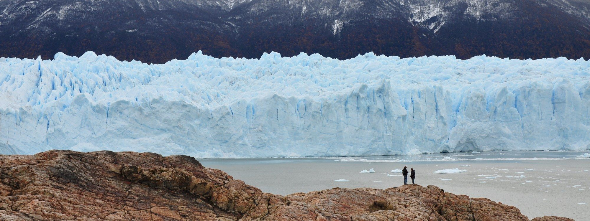 La pared de hielo azul y dentada del Glaciar Perito Moreno elevándose sobre el Lago Argentino, con dos visitantes de pie sobre las rocas en primer plano para dar escala
