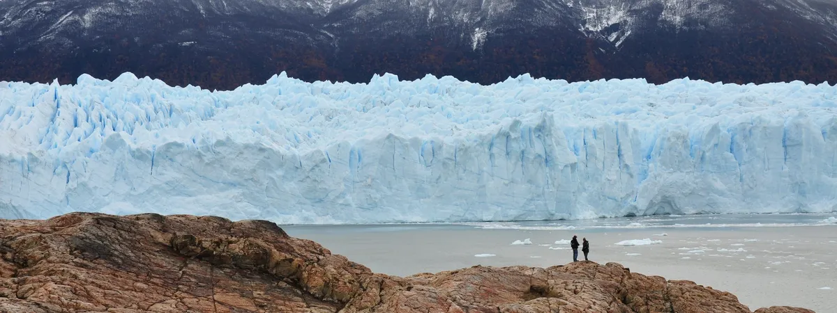 La pared de hielo azul y dentada del Glaciar Perito Moreno elevándose sobre el Lago Argentino, con dos visitantes de pie sobre las rocas en primer plano para dar escala