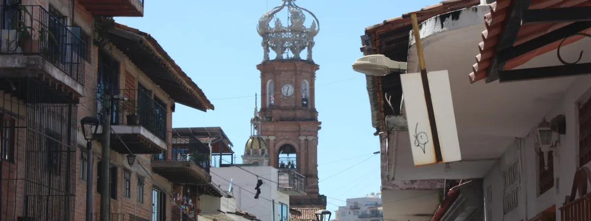 The brick bell tower and wrought-iron crown of the Parish of Our Lady of Guadalupe, framed by the balconies and rooftops of a narrow downtown street in Puerto Vallarta