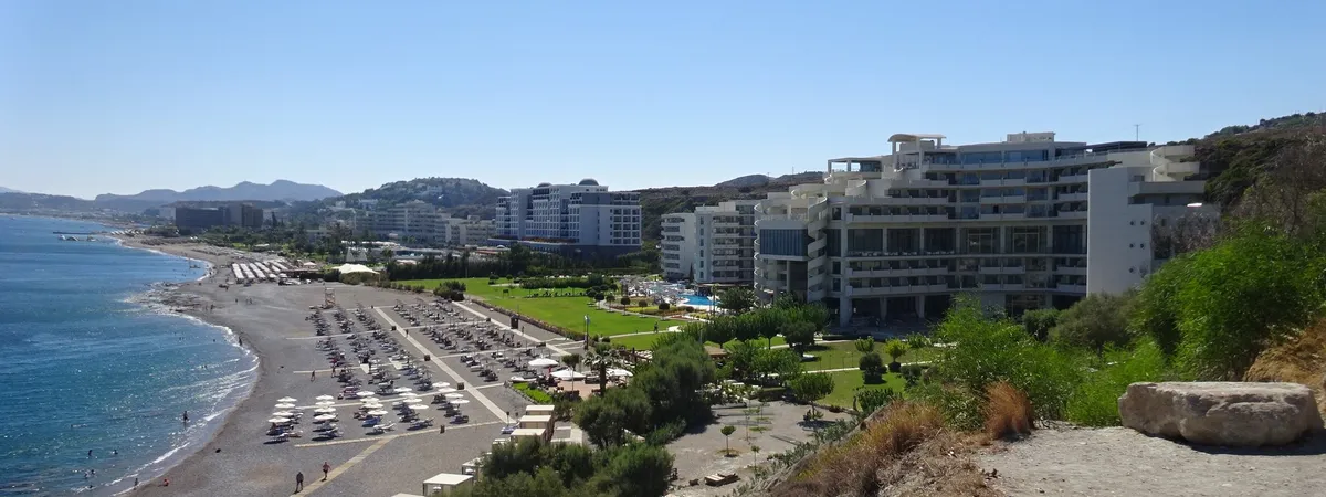 Faliraki's beachfront hotel strip along the sandy coast, seen from above with the sea and hills behind
