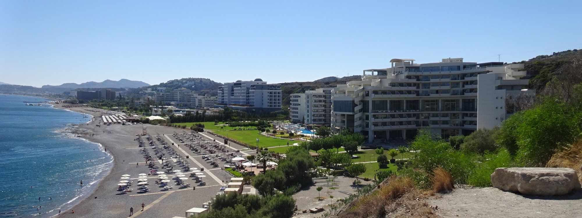 Faliraki's beachfront hotel strip along the sandy coast, seen from above with the sea and hills behind