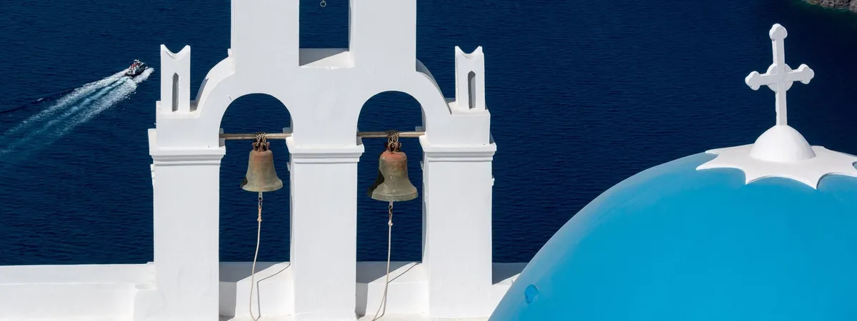 The Three Bells of Fira bell tower and a blue dome overlooking the caldera, with a boat passing below