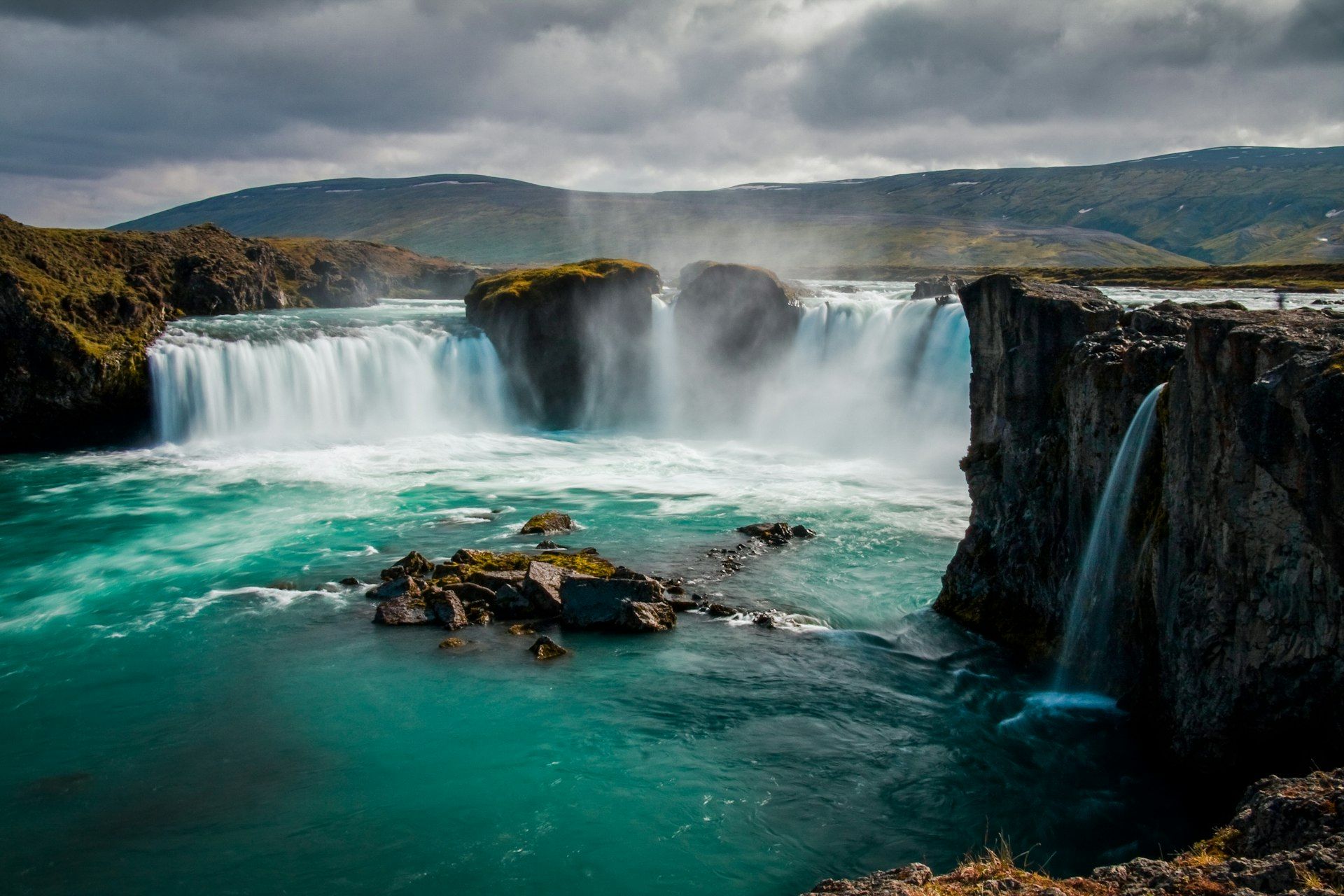 Goðafoss's horseshoe-shaped curtain of falls pouring into a turquoise pool below moss-topped rock pillars
