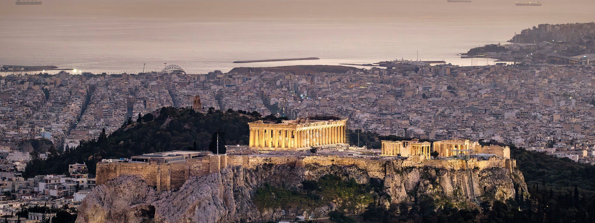 The Parthenon atop the Acropolis of Athens, lit at dusk with the city and the sea beyond