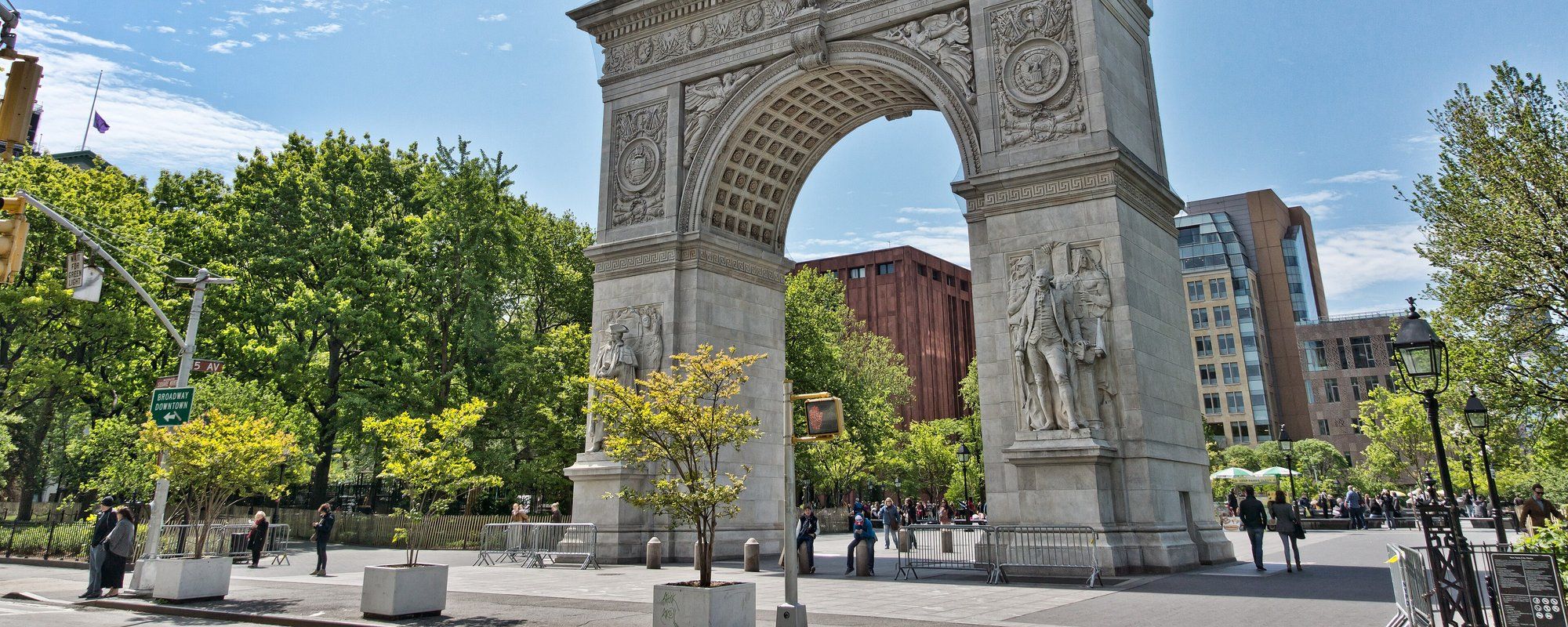 The Washington Square Arch on a sunny day, with trees and pedestrians in Washington Square Park around it