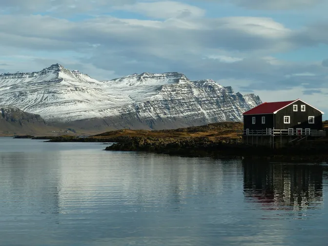 A lone house on the water's edge near Höfn, snow-streaked mountains rising behind it