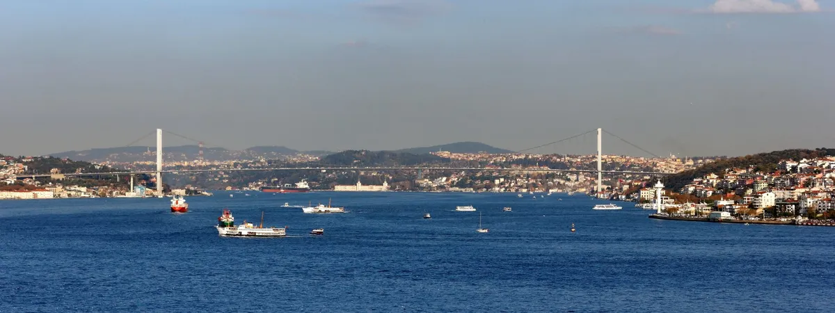 The Bosphorus Bridge connecting Europe and Asia, seen from the water with ferries crossing the strait and Istanbul's hillside neighborhoods on both shores