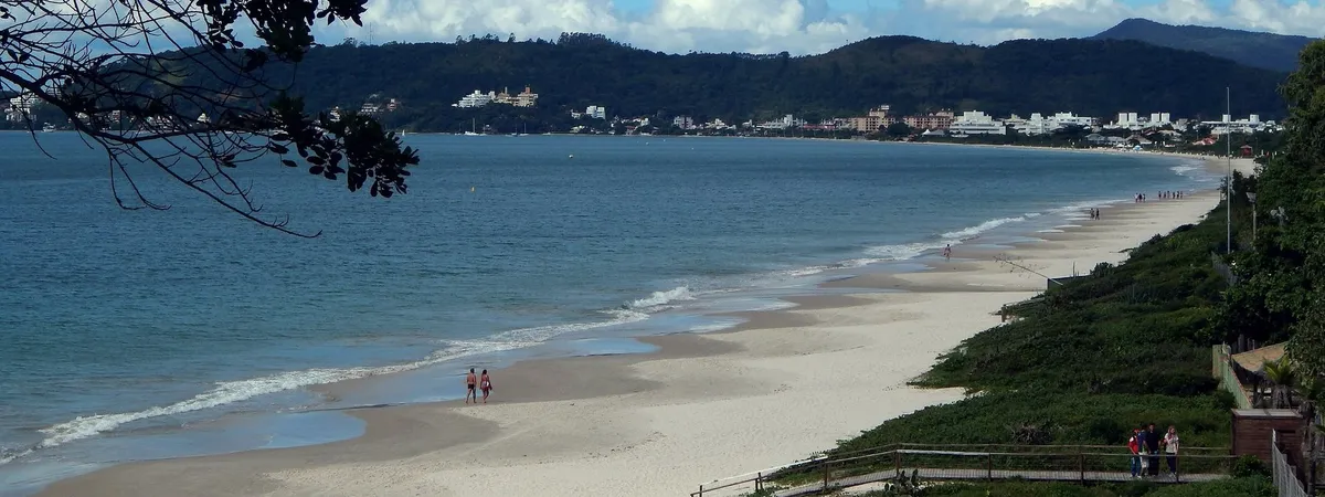 A wide sandy beach curving along the bay at Jurerê Internacional, with the town and forested hills visible in the distance