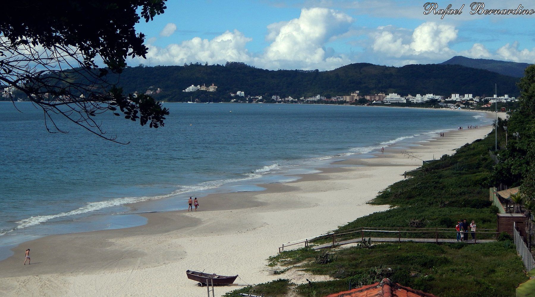 A wide sandy beach curving along the bay at Jurerê Internacional, with the town and forested hills visible in the distance