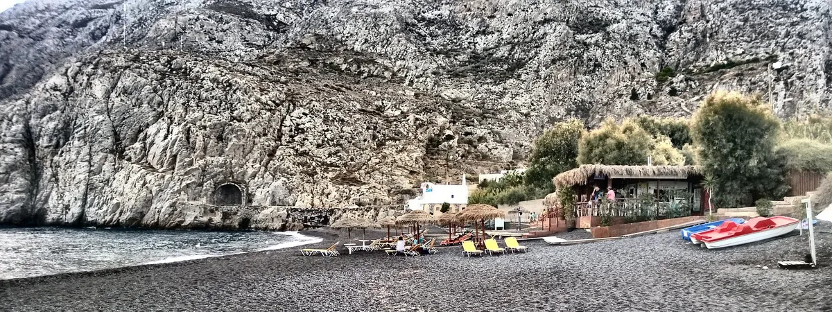 Kamari's black sand beach with sun loungers, straw umbrellas, and a beachfront taverna beneath a rocky cliff