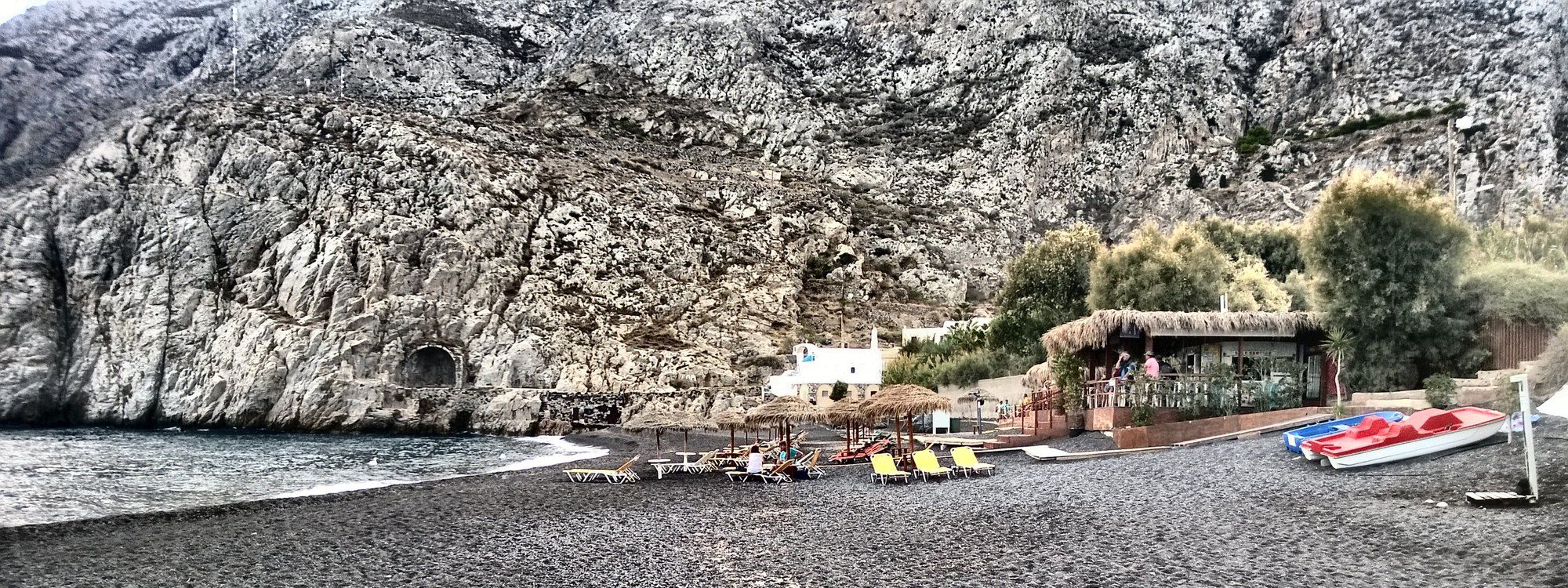 Kamari's black sand beach with sun loungers, straw umbrellas, and a beachfront taverna beneath a rocky cliff