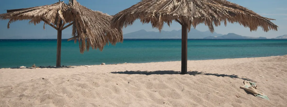 Una sombrilla de playa con techo de paja sobre la orilla de arena de Kardamena, Kos, con colinas al otro lado del agua