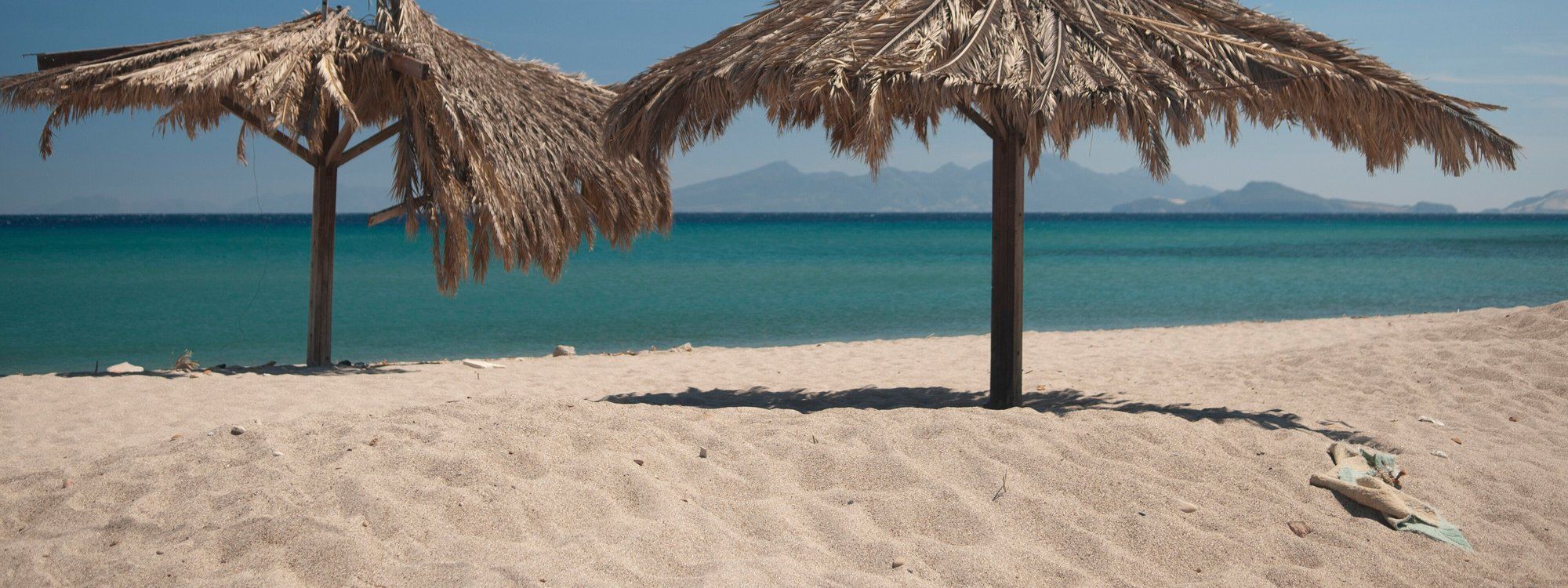 A thatched beach umbrella over the sandy shore at Kardamena, Kos, with hills across the water