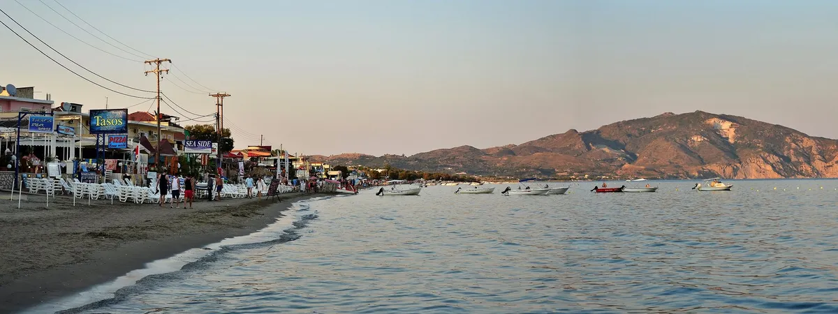 La playa de Laganas al atardecer, con tabernas y hamacas a lo largo de la arena y barcos fondeados frente a la costa