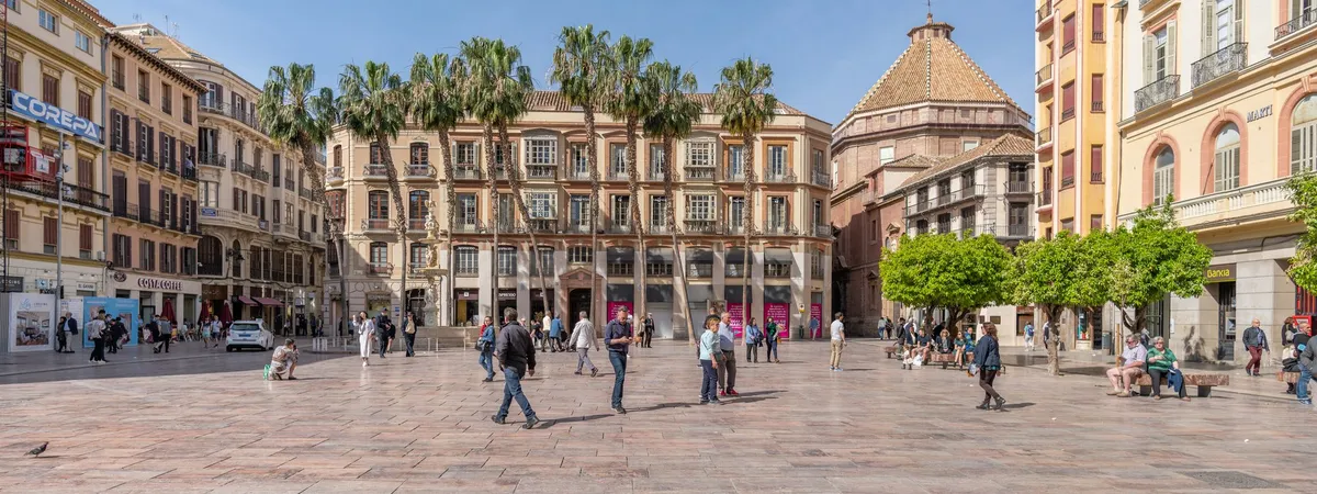 Plaza de la Constitución in Málaga's historic center, with palm trees, the Palacio Episcopal's dome, and pedestrians crossing the square on a sunny day