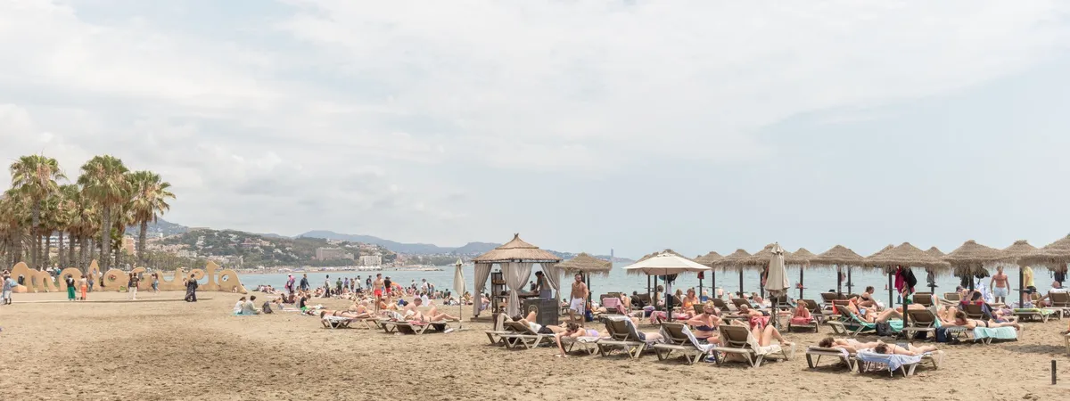 Playa de la Malagueta beach in Málaga, with the neighborhood's large sculpted sign, palm trees, sunloungers, and thatched-roof beach umbrellas along the sand