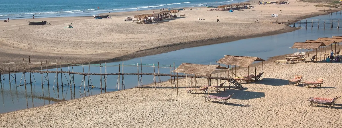 Un puente peatonal de madera que cruza la desembocadura del río en la playa de Mandrem, con chiringuitos y el mar al fondo