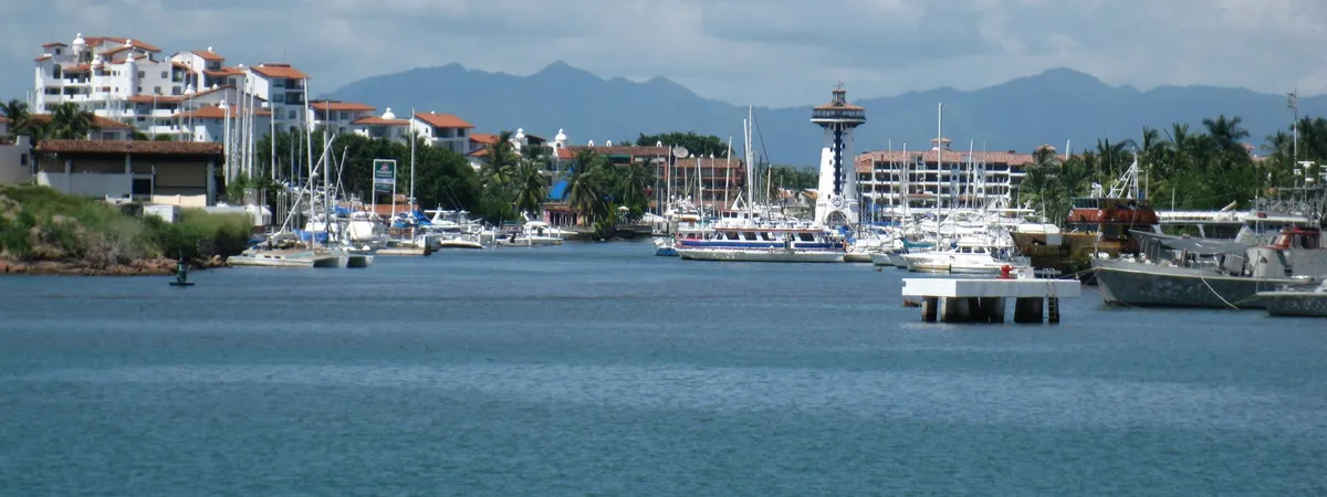 Sailboats and yachts docked along Marina Vallarta's boardwalk, with the striped El Faro lighthouse and waterfront condominiums behind, and the Sierra Madre mountains in the distance