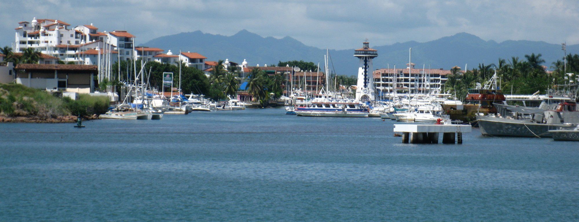 Sailboats and yachts docked along Marina Vallarta's boardwalk, with the striped El Faro lighthouse and waterfront condominiums behind, and the Sierra Madre mountains in the distance