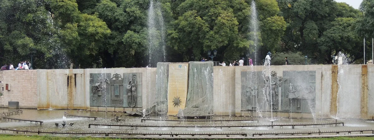 Una fuente y murales en relieve de bronce a lo largo del muro de piedra de la Plaza Independencia en el centro de Mendoza, con árboles altos detrás