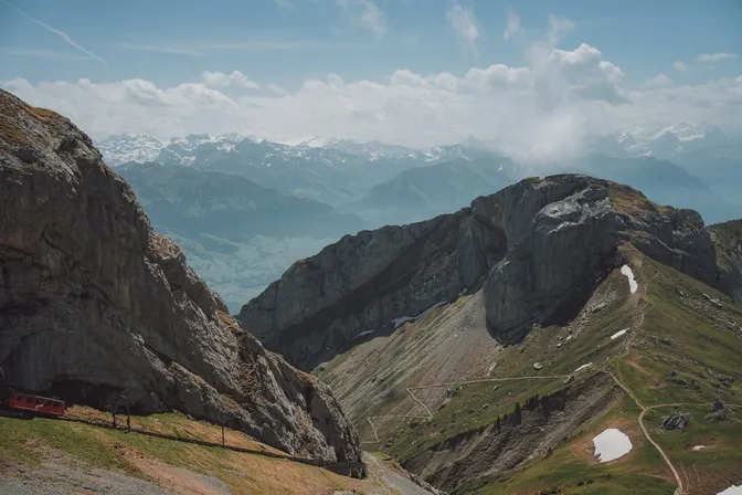 A red cogwheel train at the summit station on Mount Pilatus, with the mountain ridges falling away and the Alps beyond