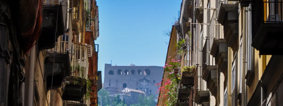 A narrow street in Naples' historic center lined with tall apartment buildings and balconies, opening onto a view of a domed monastery building in the distance