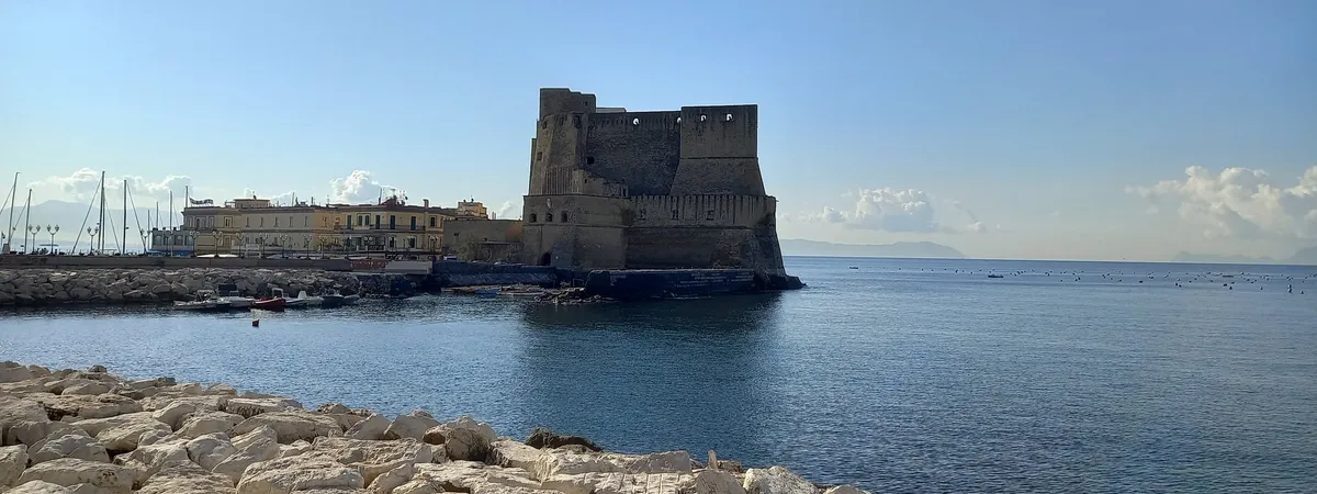 Castel dell'Ovo, a medieval waterfront fortress in Naples, seen across the harbor with the marina and hilltop city behind it