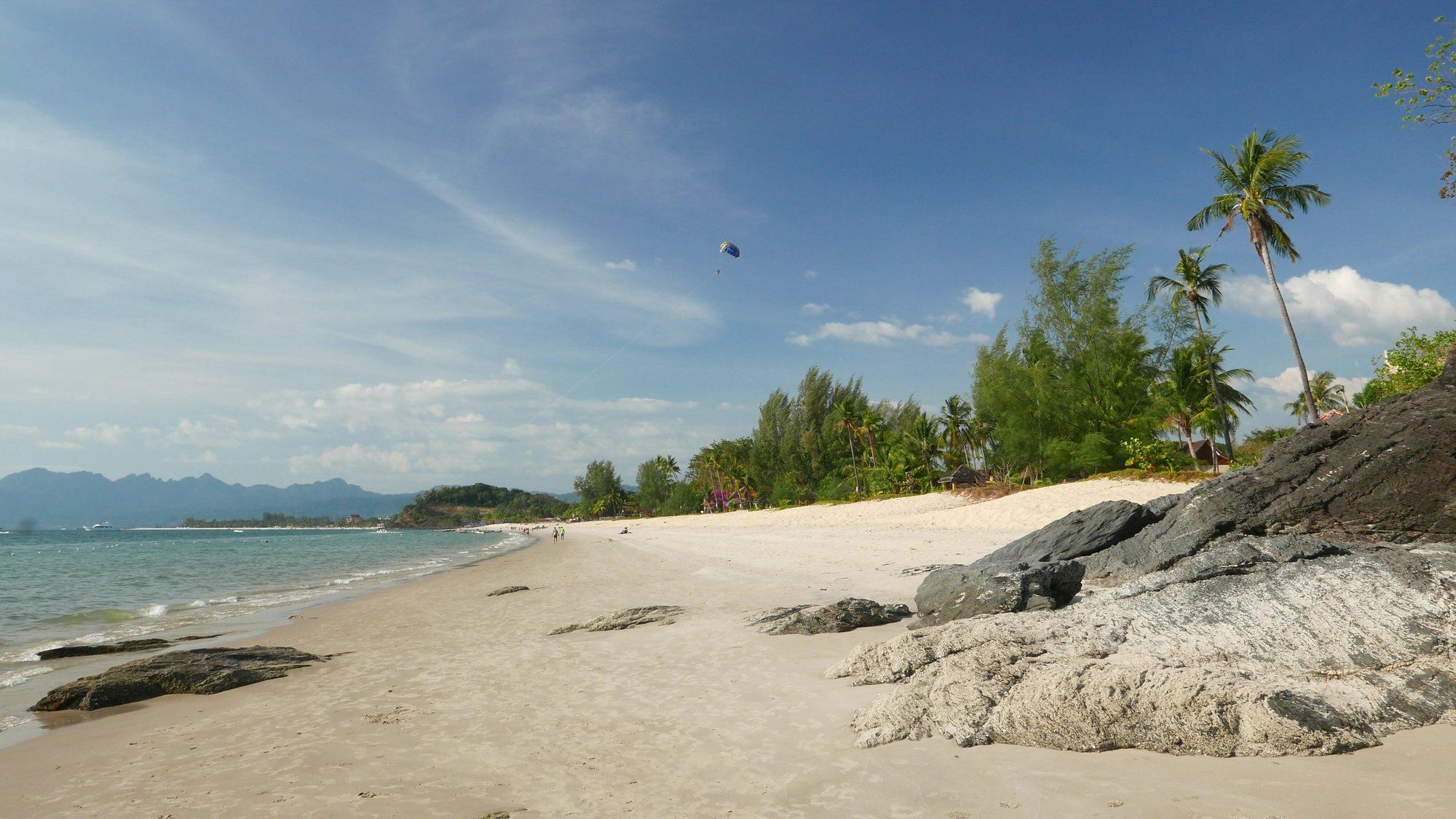 The white sand and dark rock outcrops of Cenang Beach in Langkawi, with a parasailer overhead