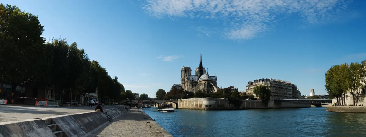 Notre-Dame Cathedral seen across the Seine from the riverside walkway, with the Île de la Cité and a stone bridge behind it