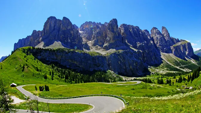 Hairpin bends on the Passo Gardena road curving through summer meadows, with a wall of Dolomite towers behind